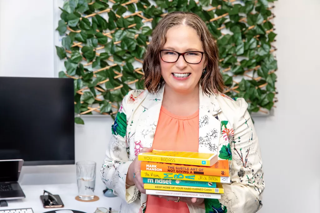 Anastasia Geneave from No Grey Suits holding a stack of books, including The Visual MBA, The Confessions of a Misfit Entrepreneur and You Are A Badass. Image is for her Copywriting Packages webpage.