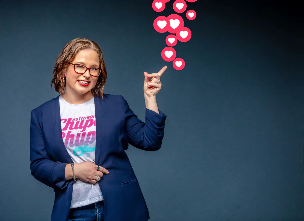 ALT text for accessibility: The image features Anastasia Geneave from No Grey Suits, smiling and pointing upwards at a cascade of pink heart icons that seem to emanate from her finger, symbolizing social media 'likes' or 'love' reactions. She is wearing glasses, a white t-shirt with the colorful #STAYBRIGHT Chupa Chups logo, and a dark blazer. Her friendly demeanor, paired with professional attire, projects a positive and approachable image in a social media management context.