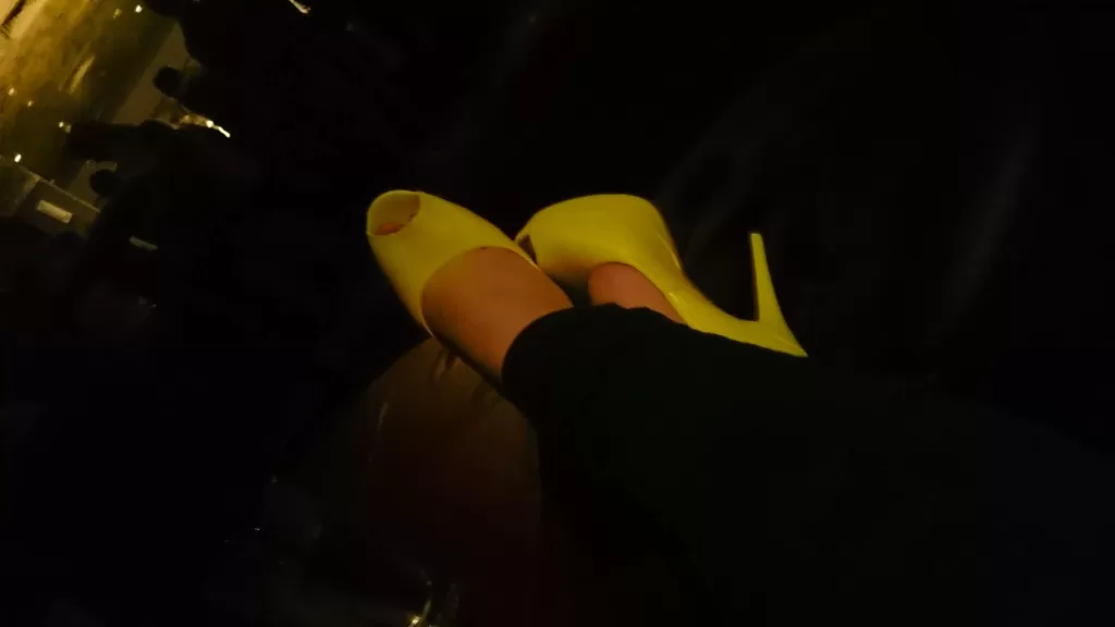 The image displays a pair of bright yellow, high-heeled, open-toe shoes being worn. The setting appears to be dimly lit, with ambient light creating a soft glow on the shoes. The wearer's feet are crossed at the ankles, suggesting a casual yet elegant posture. The high heels are the focal point of the image, emphasized by the contrast with the darker surroundings, likely indicating a sense of style or fashion statement.